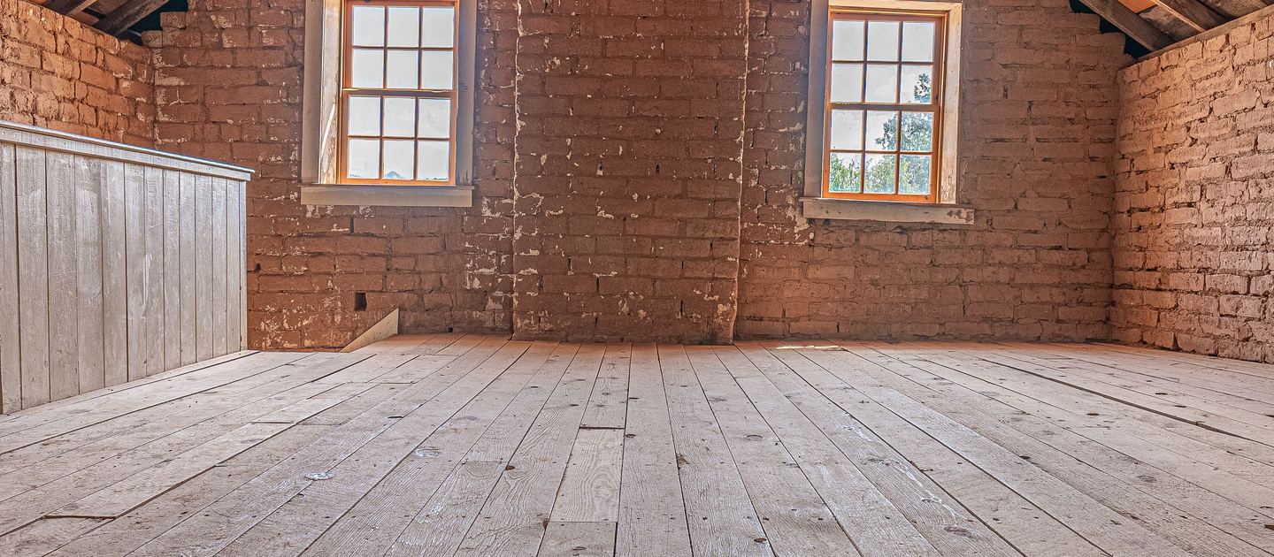 An empty loft with a bare timber floor, a brick gable wall and two windows.